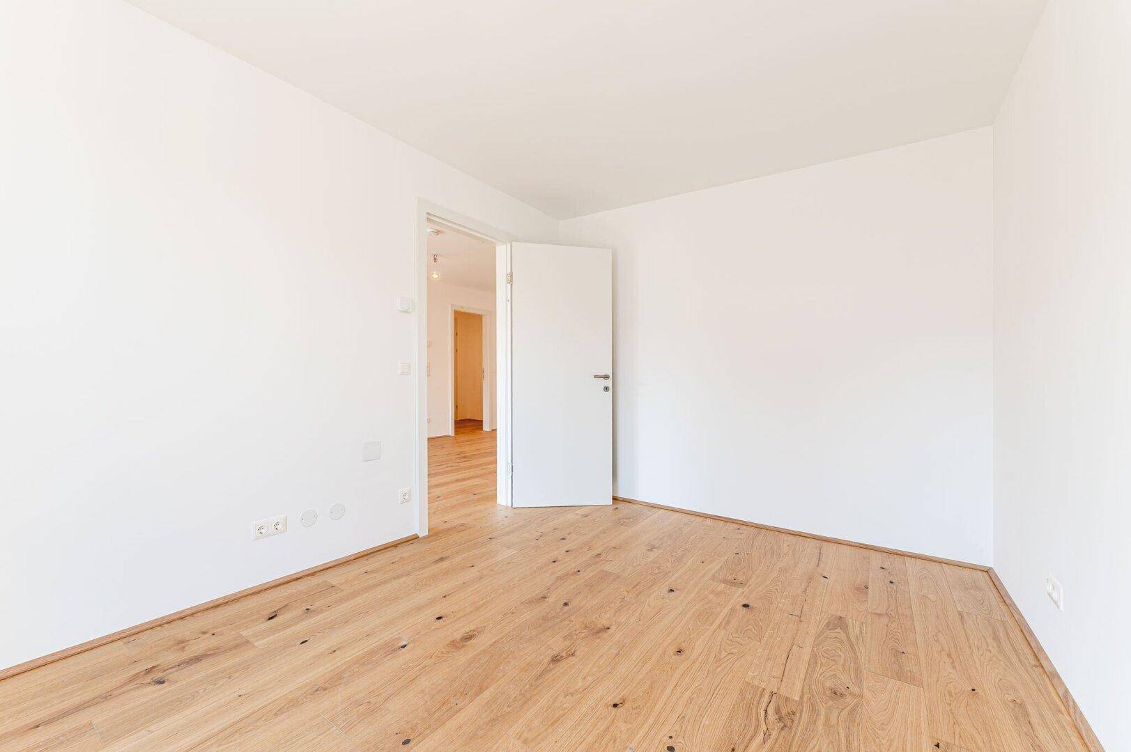Leeres, helles Zimmer mit Holzboden und weiß gestrichenen Wänden, ideal für Schlaf- oder Arbeitszimmer.