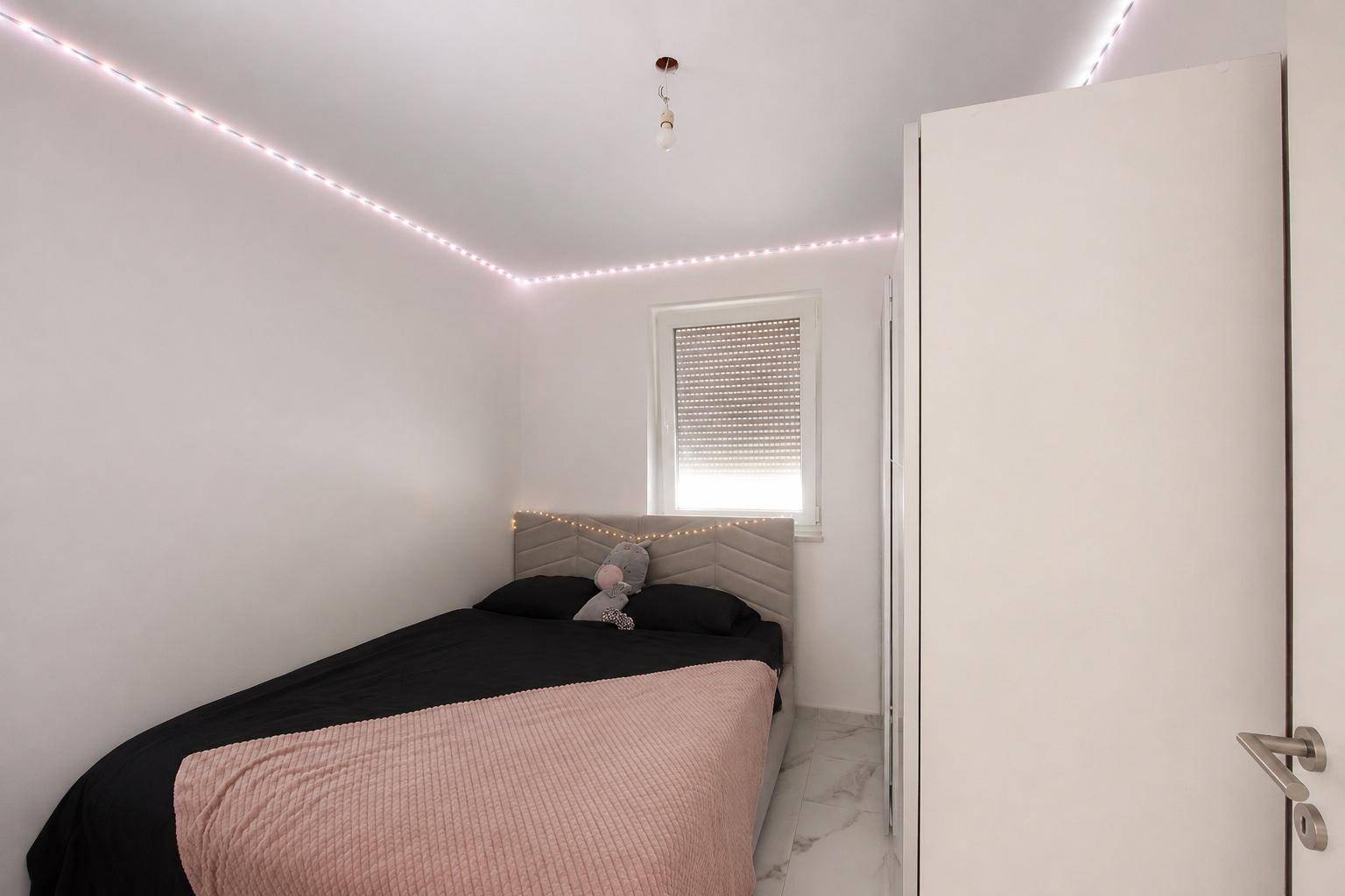 Helles Schlafzimmer mit Einzelbett, Fenster und indirekter Deckenbeleuchtung.