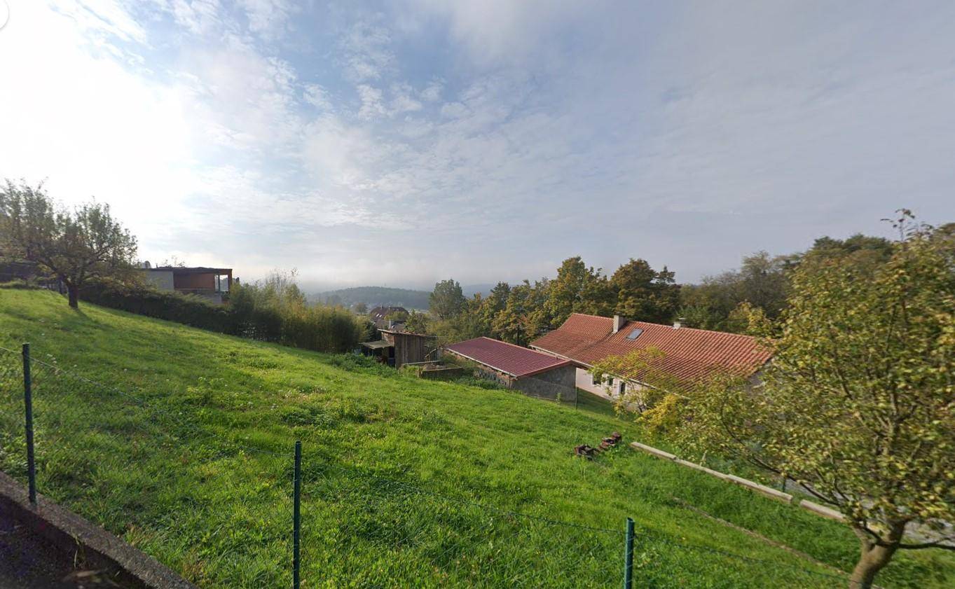 Panoramablick über ein grünes Grundstück mit einem Haus und bewaldeter Landschaft unter bewölktem Himmel.