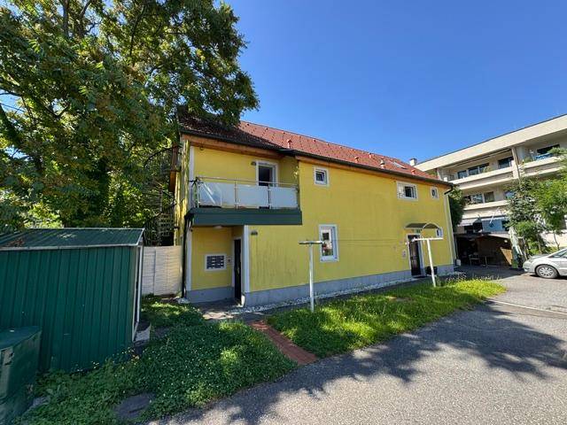 Seitenansicht des gelben Gebäudes mit Balkon, grünem Schuppen und Rasenfläche im Vordergrund.