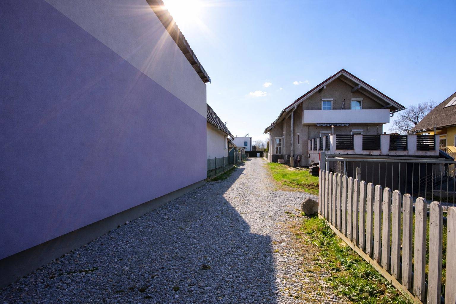 Kiesweg zwischen zwei Wohnhäusern mit unterschiedlichen Fassadenfarben unter blauem Himmel.