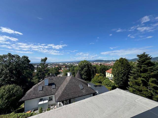 Panoramablick von der Dachterrasse über die Stadt und die umliegende grüne Landschaft unter blauem Himmel.