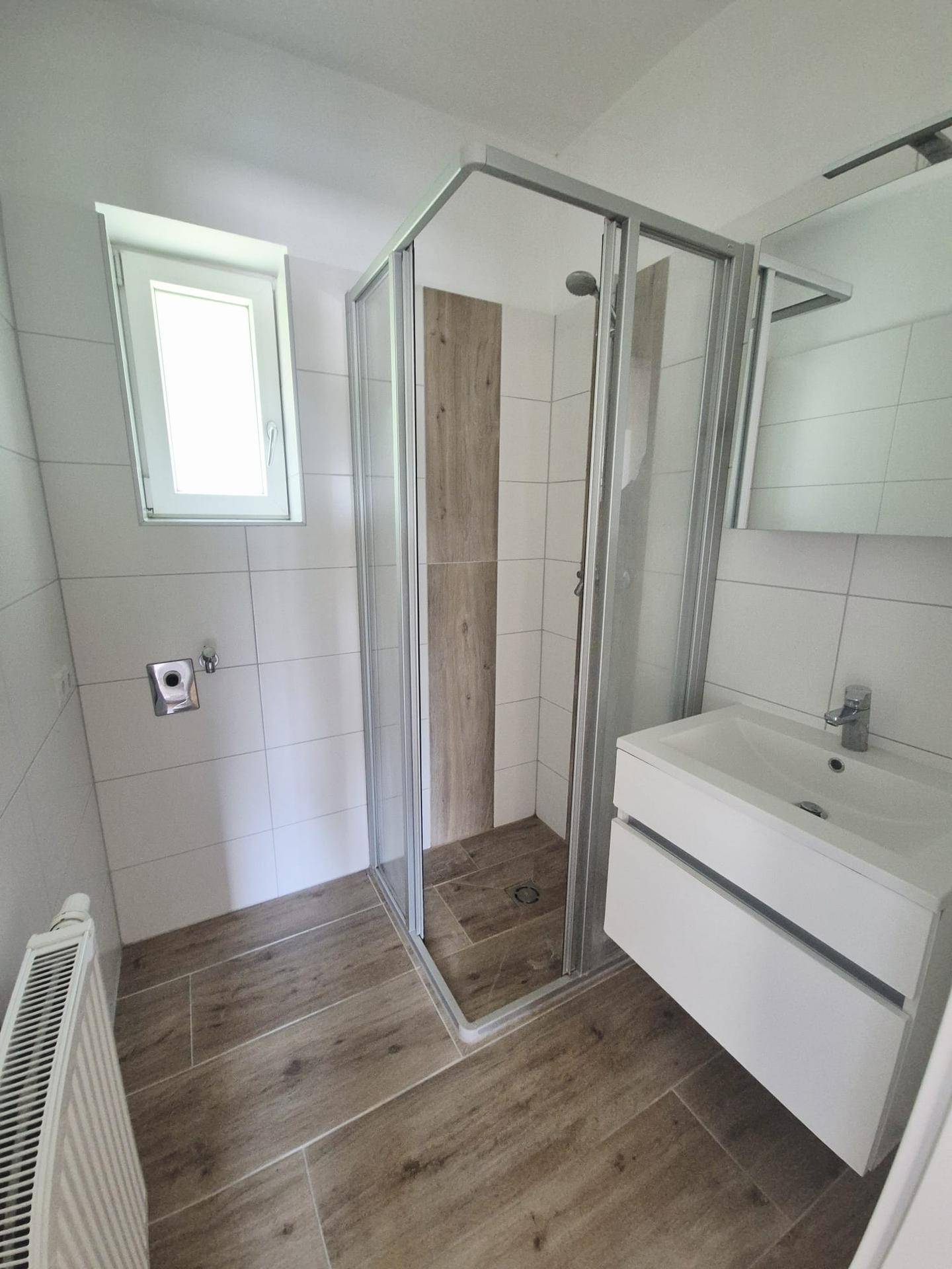 Modernes Badezimmer mit Eckdusche, Waschtisch und Fenster, hell gefliest mit Holzoptik-Boden.