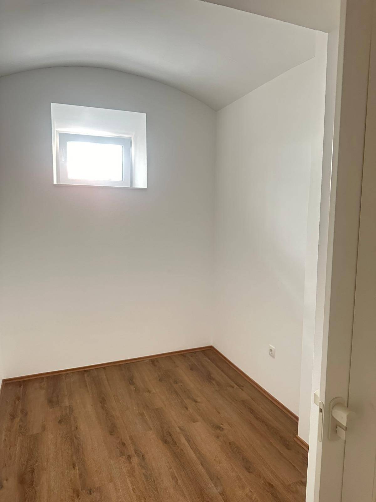 Helles Zimmer mit Laminatboden und einem kleinen Fenster, ideal als Schlaf- oder Arbeitszimmer.