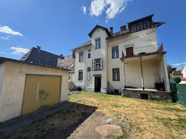Rückansicht des historischen Gebäudes mit einer Garage, einem kleinen Balkon und einem verwilderten Gartenbereich.
