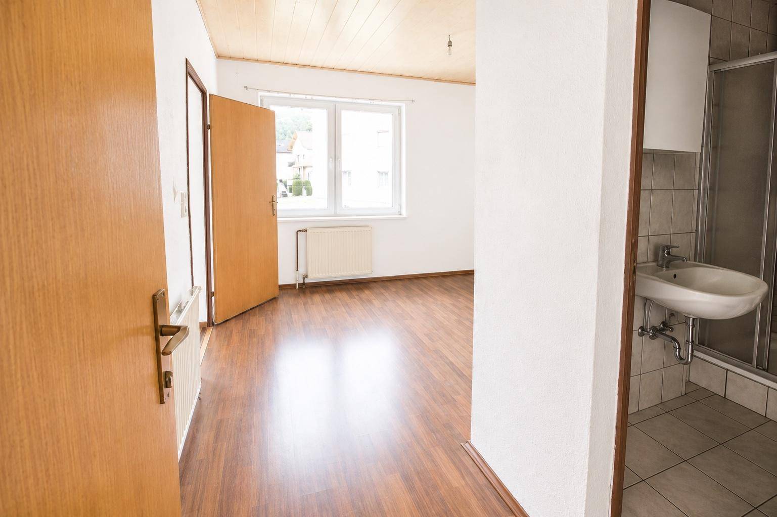 Raum mit Laminatboden und Zugang zu einem kleinen Badezimmer mit Waschbecken und Dusche.