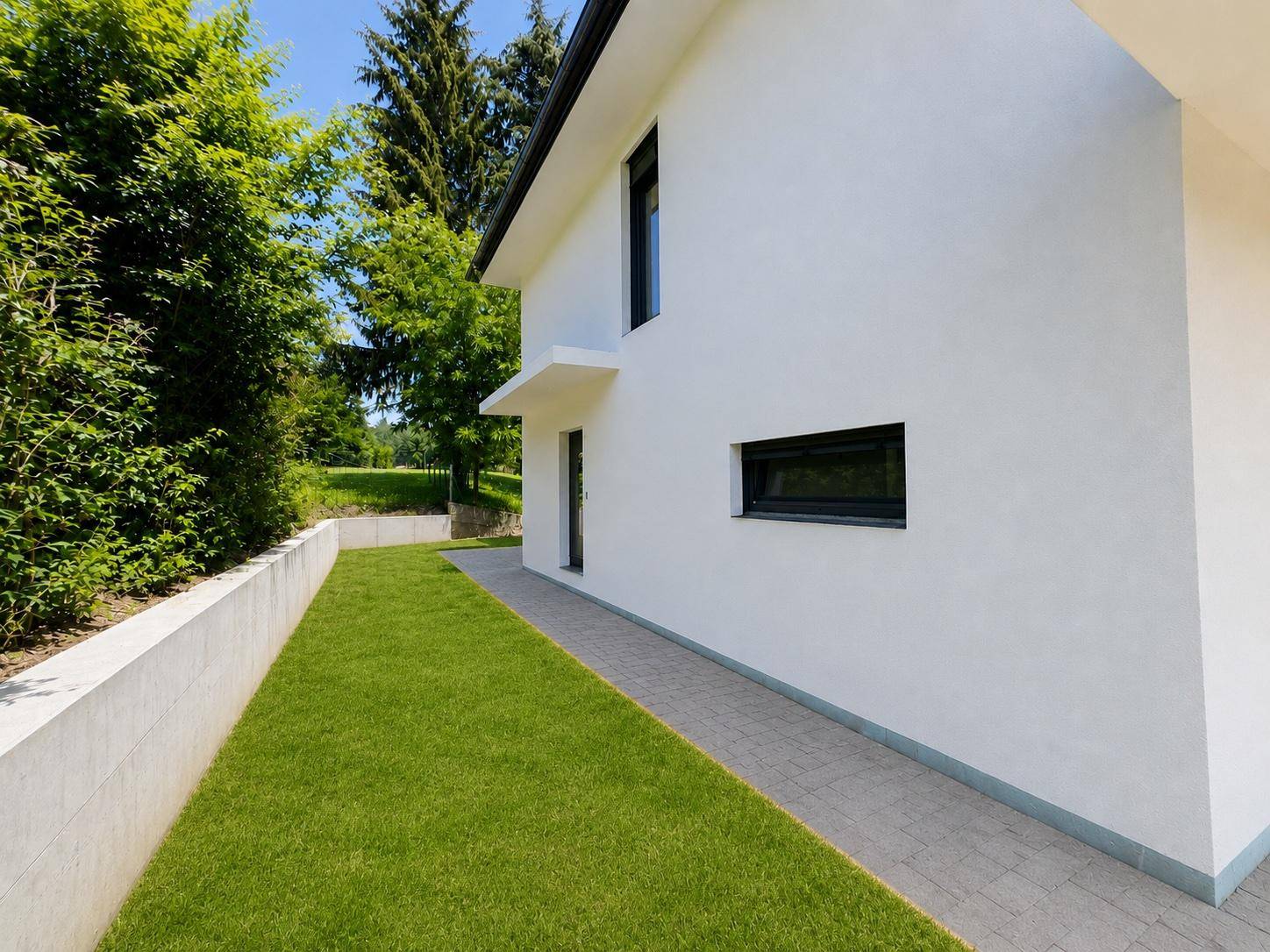 Seitlicher Weg entlang des Hauses mit Rasenfläche, Stützmauer und grüner Bepflanzung.