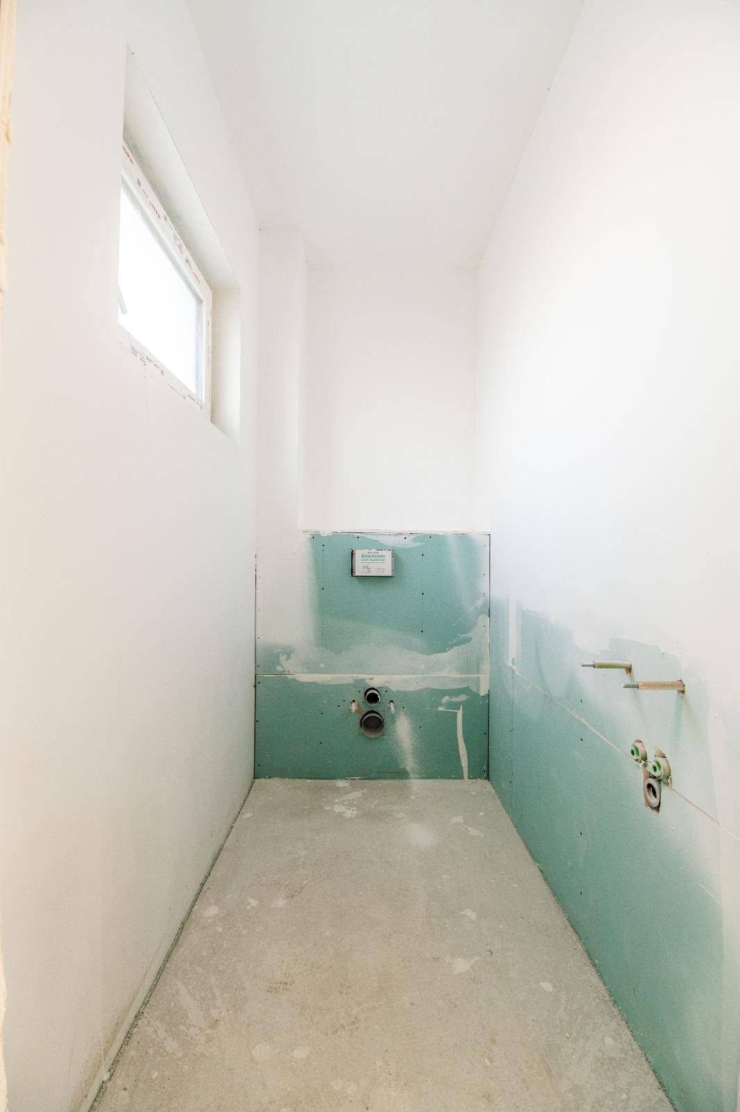 Unfertiges Gäste-WC im Rohbau mit Fenster und vorbereiteten Anschlüssen.