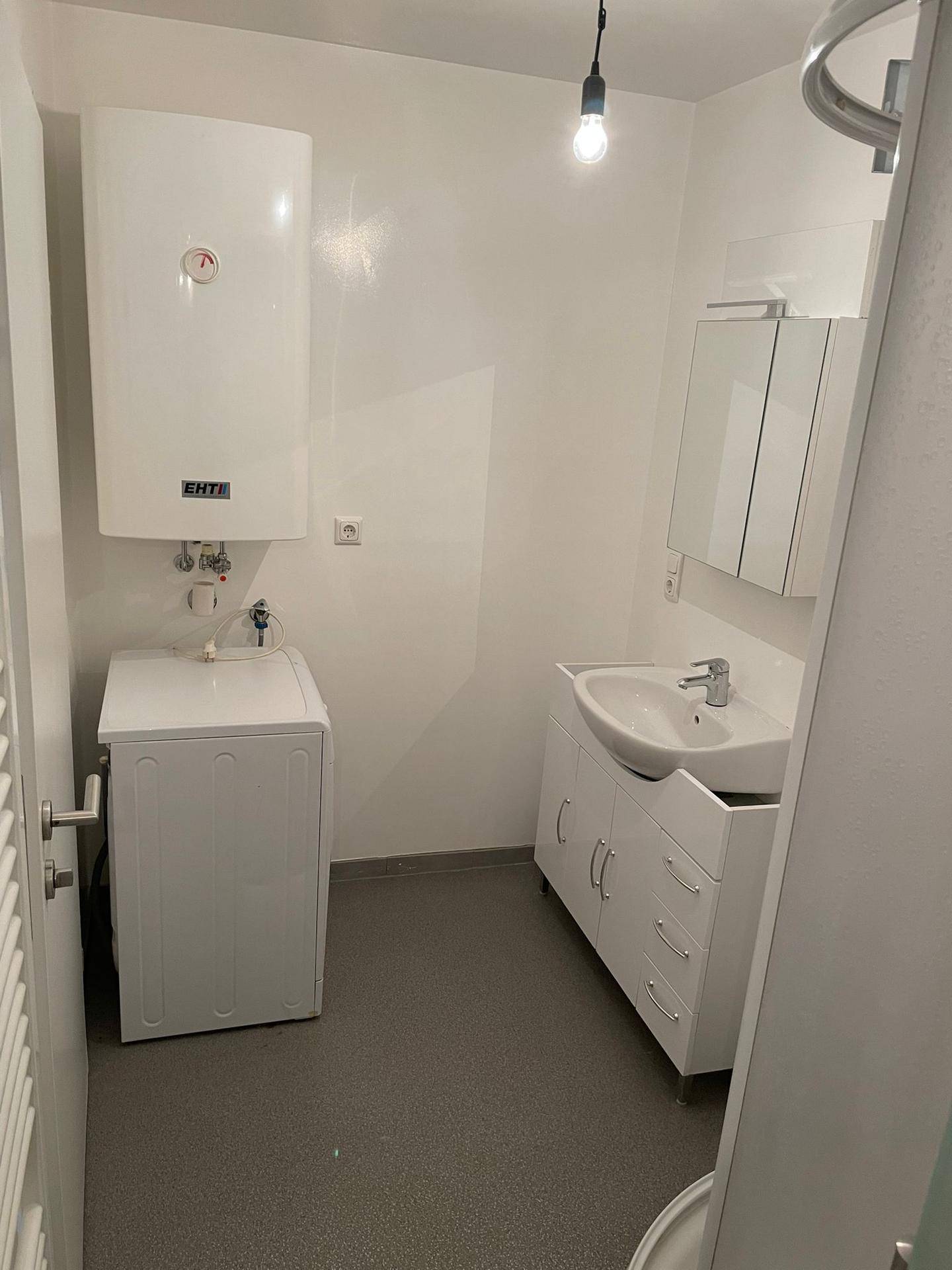 Badezimmer mit Waschmaschine, Waschbeckenunterschrank, Spiegelschrank und Warmwasserboiler.