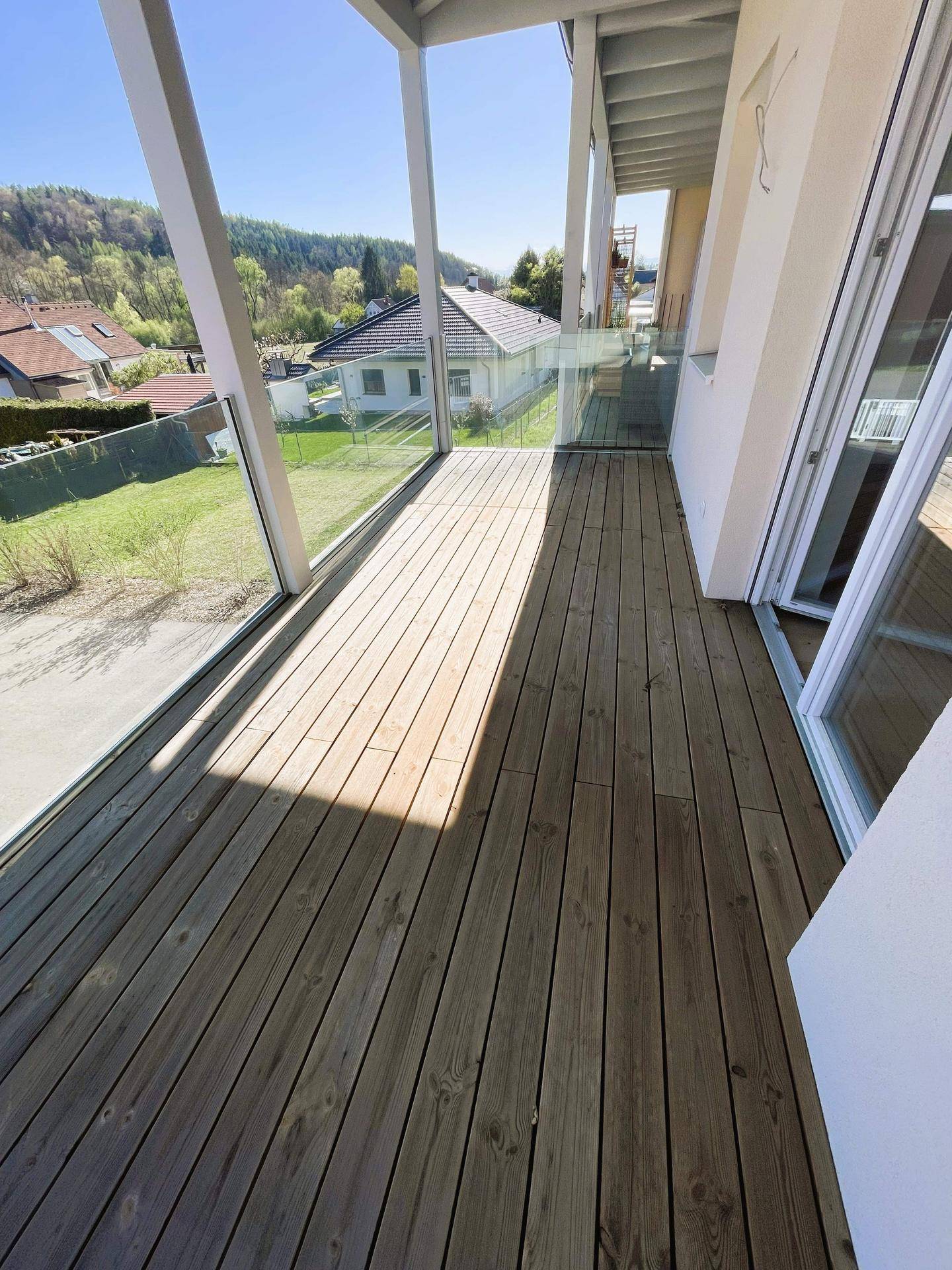 Geräumiger Balkon mit Holzboden und Glasgeländer, bietet einen schönen Blick ins Grüne.