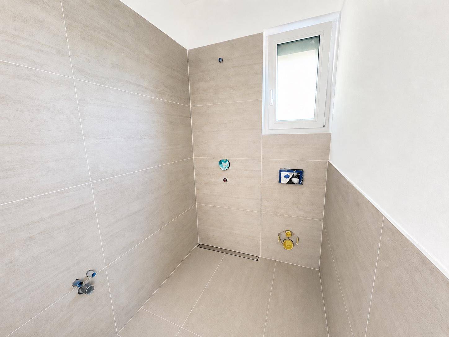 Modernes Badezimmer im Rohbau mit hellgrauen Fliesen, Fenster und vorbereiteten Anschlüssen für Dusche und WC.