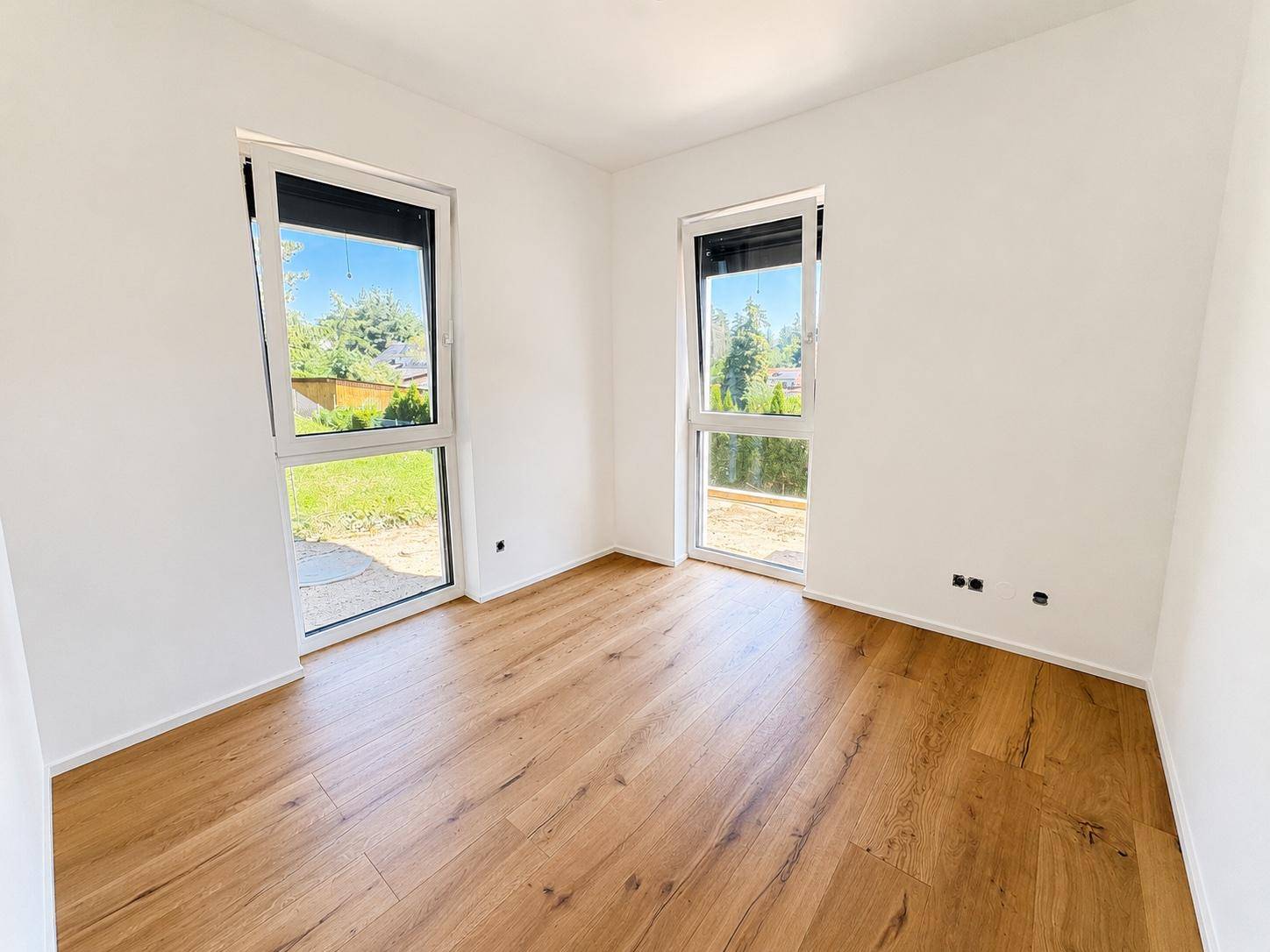 Helles Zimmer mit Holzboden, weißen Wänden und zwei großen Fenstern, die viel Licht hereinlassen.
