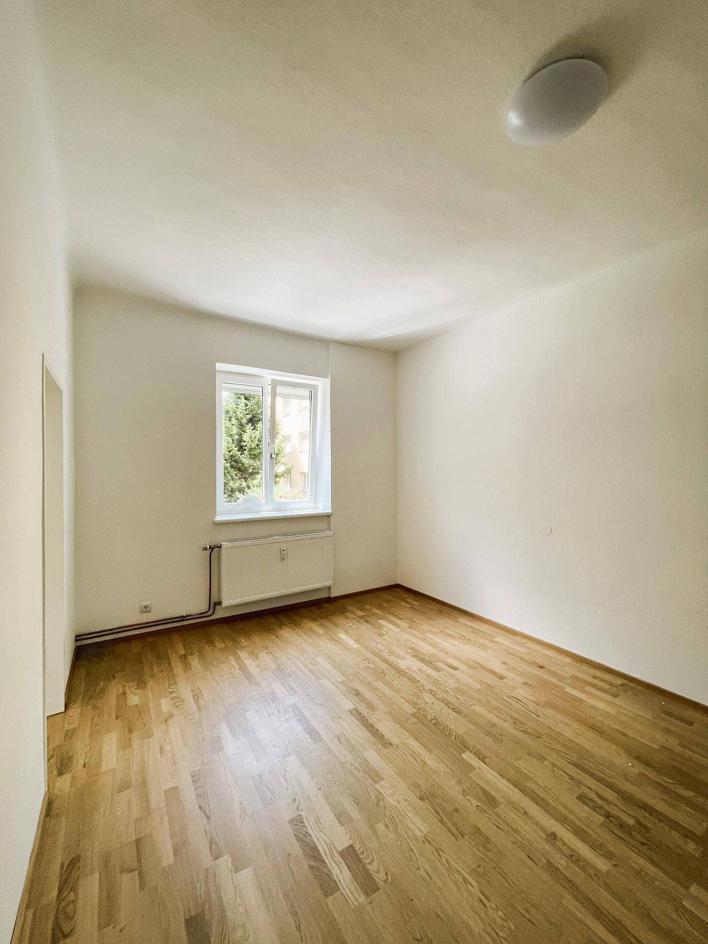 Geräumiges, helles Zimmer mit Parkettboden, weißen Wänden und Blick ins Grüne durch das Fenster.