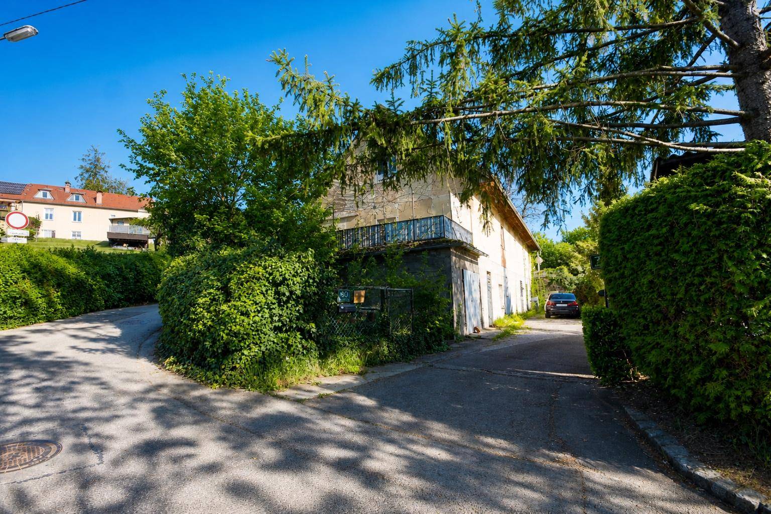 Altes Gebäude mit Balkon und üppiger Vegetation an einer Zufahrtsstraße.