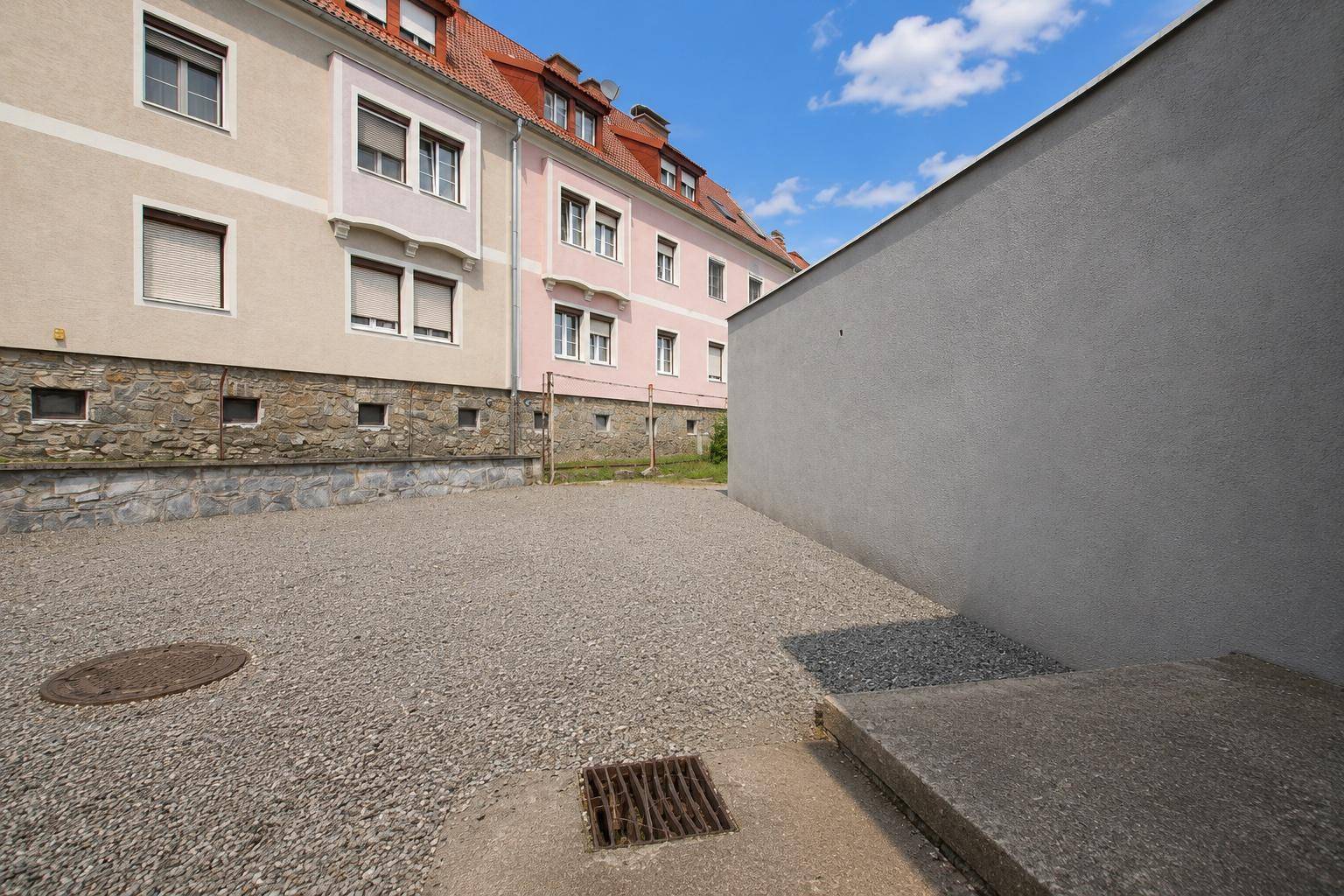Schotterfläche zwischen zwei Gebäuden mit einem Gully und einer Steinmauer im Vordergrund.