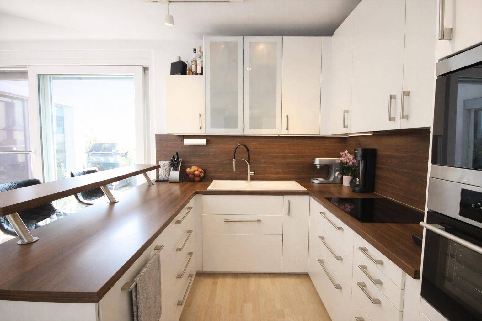 Helle Küche mit U-förmiger Anordnung, Spüle am Fenster und modernen Geräten.