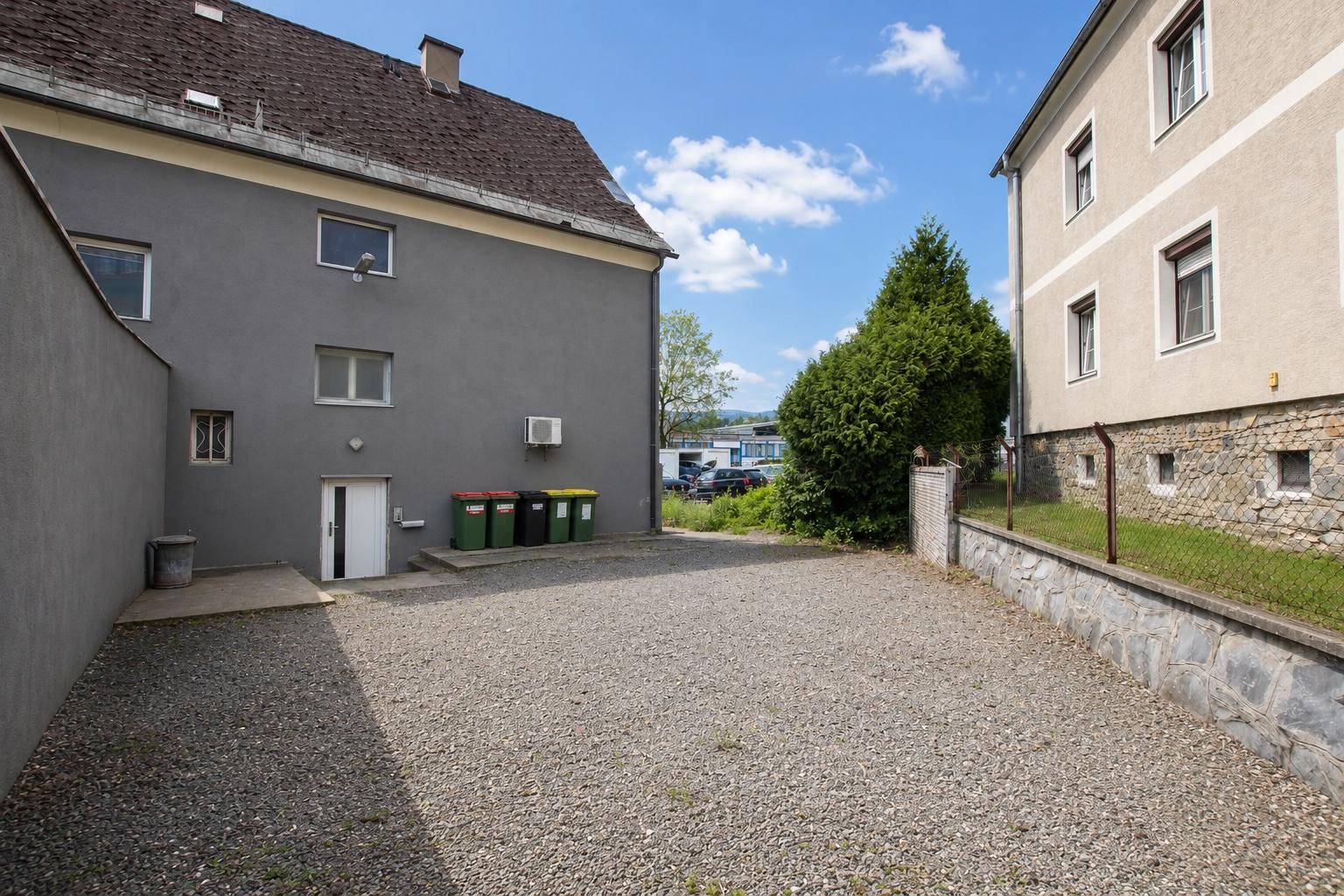 Rückseite des Gebäudes mit grauem Putz, einem Schotterparkplatz und Mülltonnen vor einem grünen Hügel.
