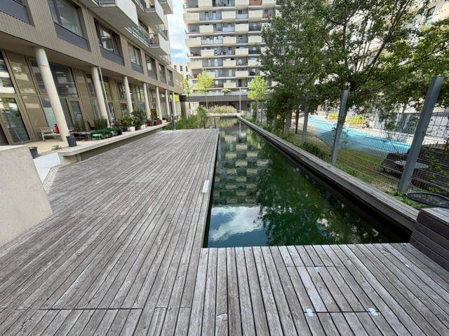 Außenbereich mit Holzdeck und einem langen Wasserbecken, umgeben von modernen Gebäuden und Bäumen.