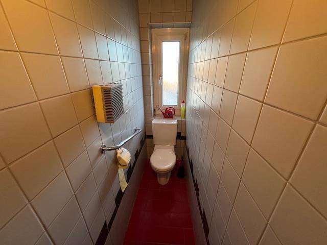 Schmale Toilette mit Fenster, Haltegriff und Fliesen an den Wänden und rotem Boden.