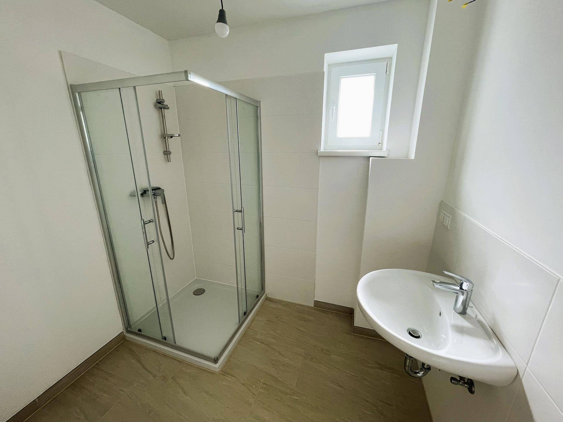 Modernes Badezimmer mit Duschkabine, Waschbecken und Fenster, hell gefliest.