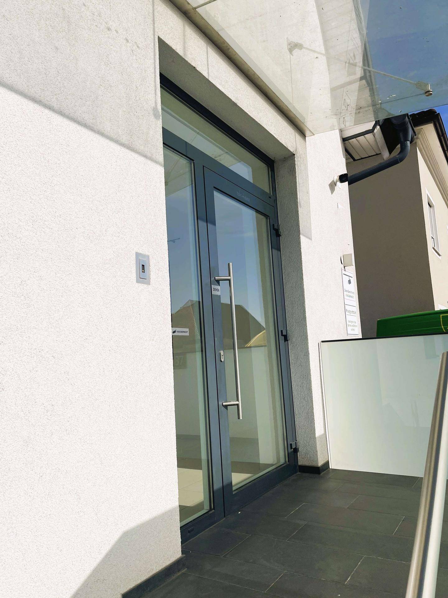 Moderner Eingangsbereich mit Glastür und Vordach, umgeben von weißer Fassade.
