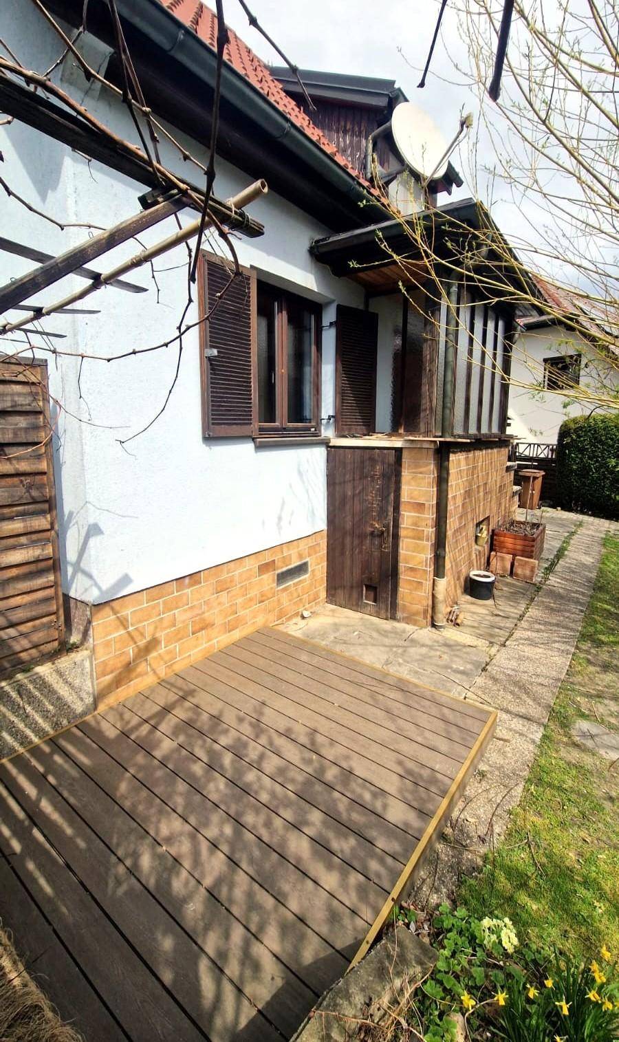Hölzerne Terrasse am Haus mit Ziegelmauerwerk, Fensterläden und einem kleinen Gartenbereich.
