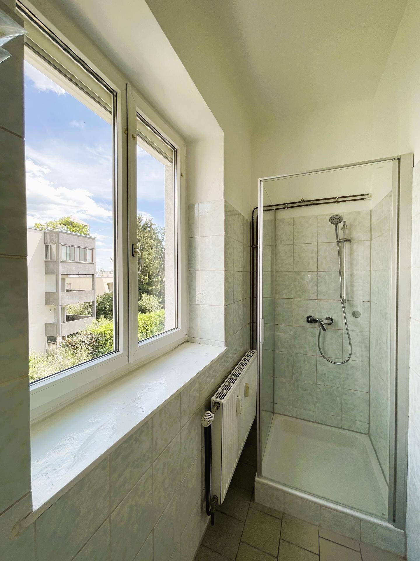 Badezimmer mit Duschkabine, hellen Fliesen und einem Fenster, das Tageslicht hereinlässt.