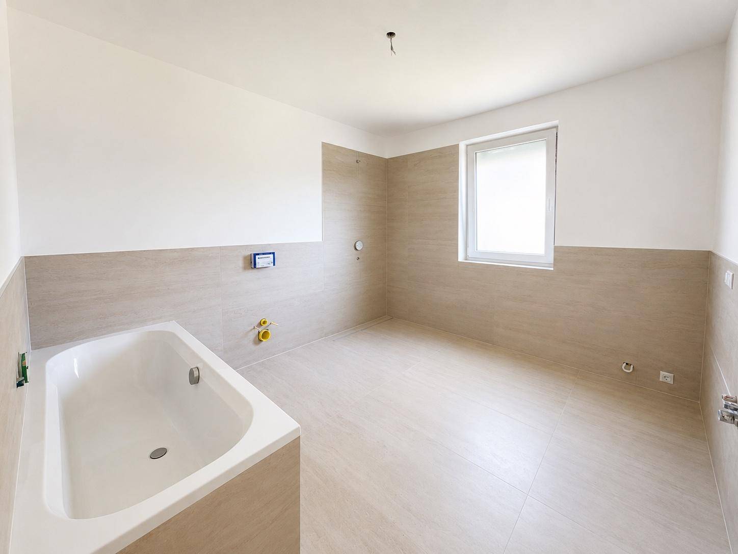 Geräumiges Badezimmer im Rohbau mit beigefarbenen Fliesen, Fenster und eingebauter Badewanne.