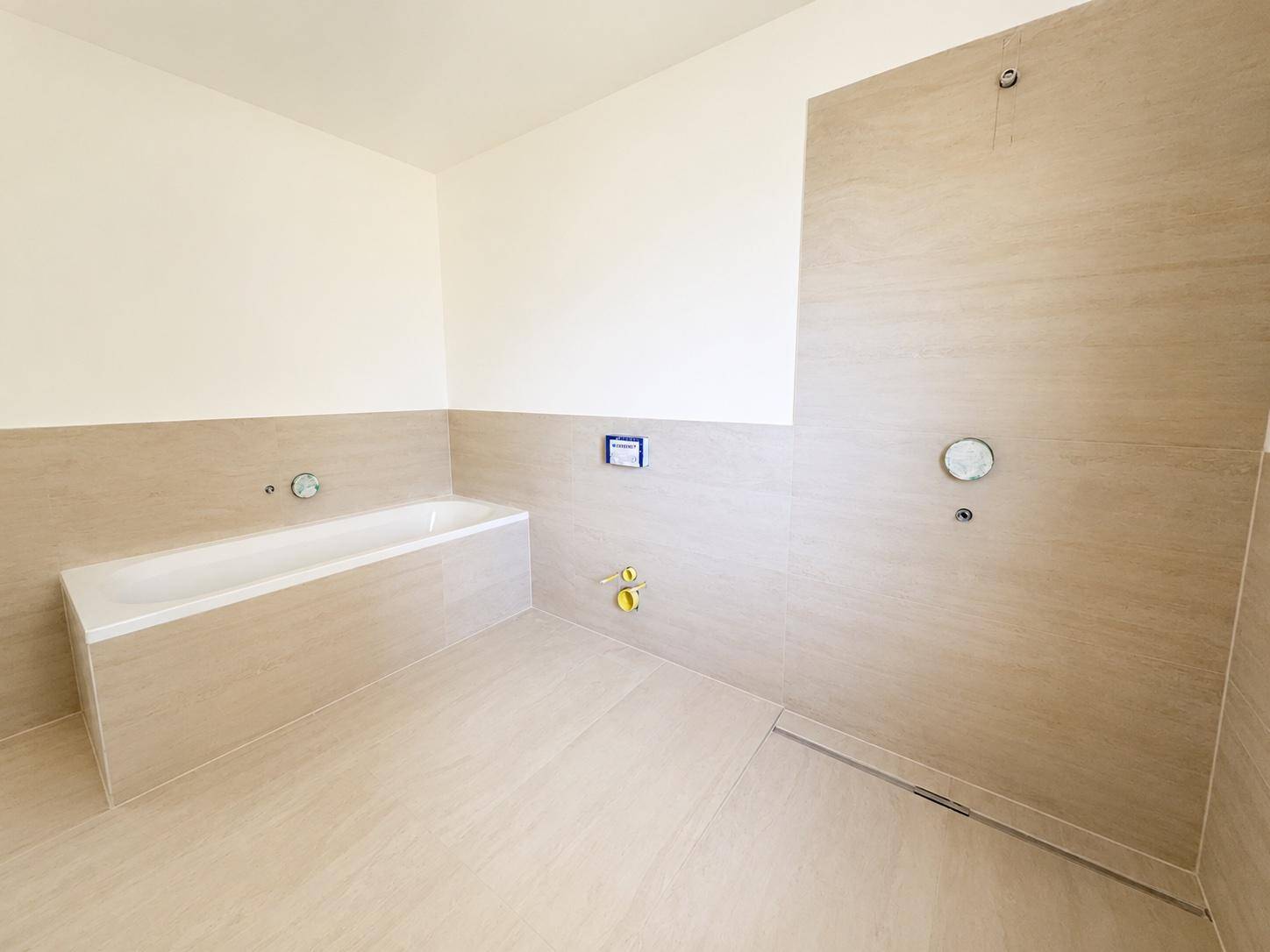 Modernes Badezimmer im Rohbau mit Badewanne, hellen Fliesen und vorbereiteten Duschanschlüssen.