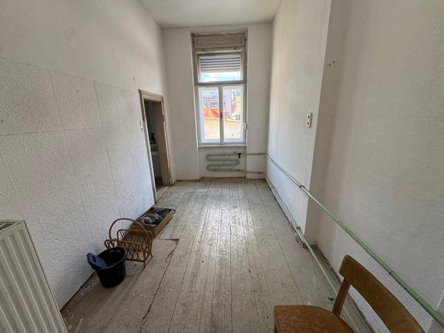 Langer, schmaler Raum mit Dielenboden und Heizkörper unter dem Fenster, renovierungsbedürftig.