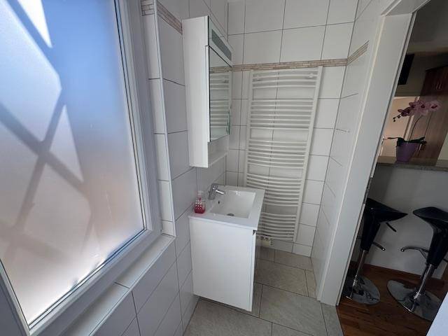 Helles Badezimmer mit Waschbecken, Spiegelschrank, Handtuchheizkörper und Fenster.