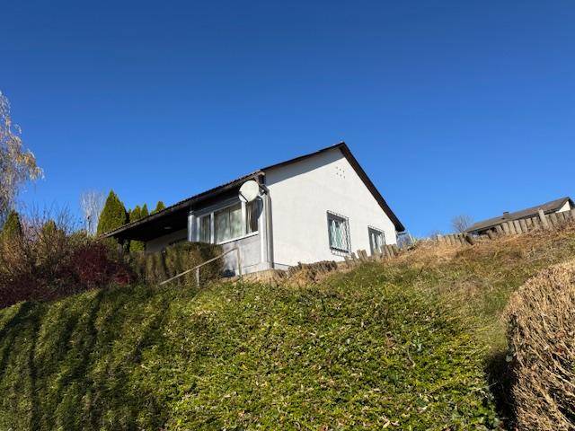 Weißes Haus auf einem Hügel, umgeben von Hecken und Bäumen unter blauem Himmel.