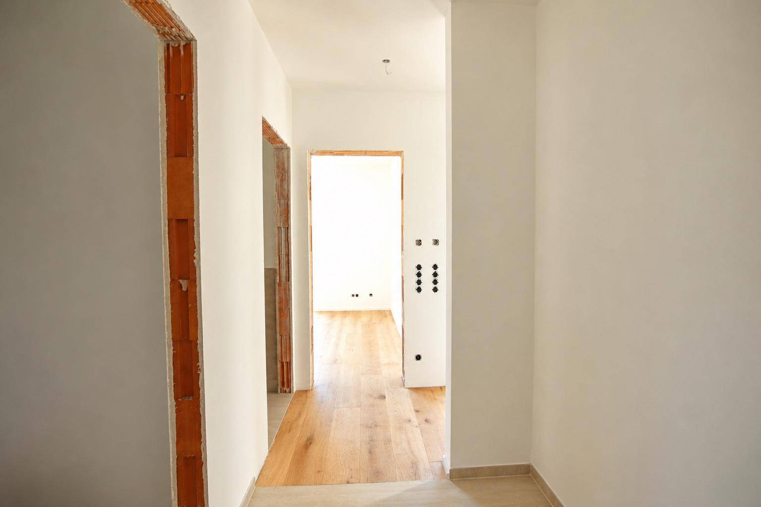 Heller Flur im Rohbau mit Holzboden und unverputzten Türrahmen, der zu weiteren Räumen führt.