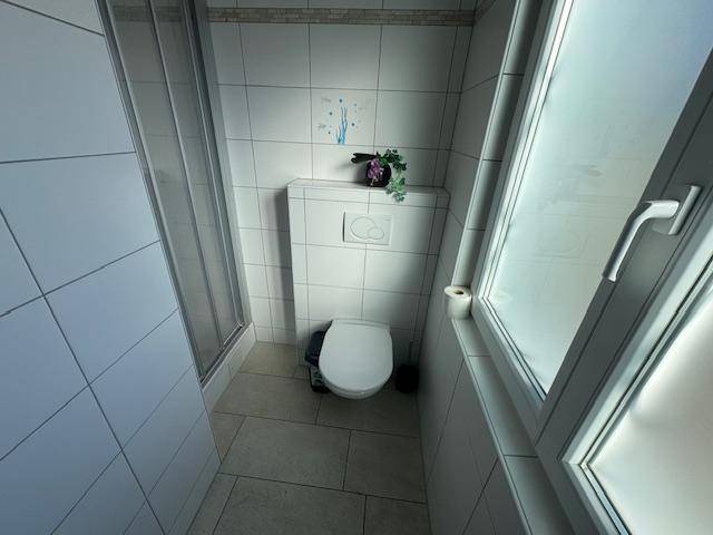 Modernes Badezimmer mit Duschkabine, WC und Fenster, hell gefliest.