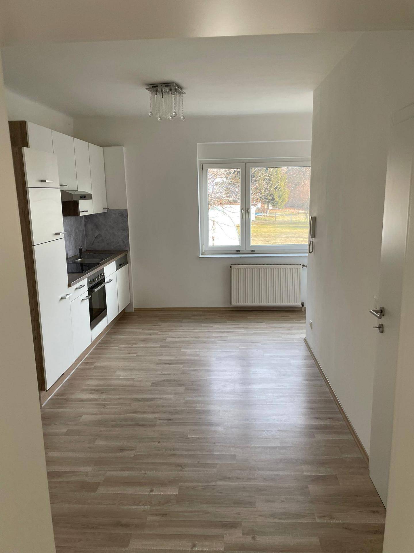 Offener Wohnbereich mit integrierter weißer Küche, Laminatboden und großem Fenster.