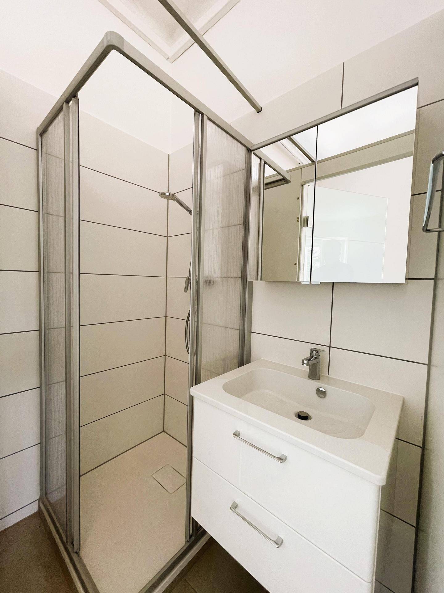 Helles Badezimmer mit Duschkabine, modernem Waschtisch und Spiegelschrank, komplett weiß gefliest.