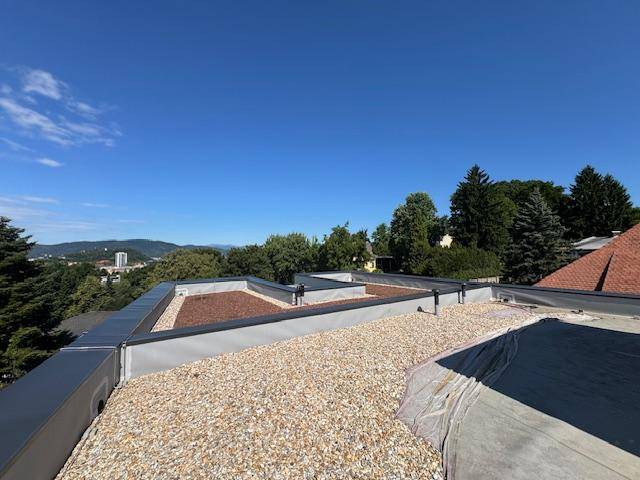 Dachfläche in Bau mit Kiesbelag und Blick auf die grüne Umgebung unter blauem Himmel.