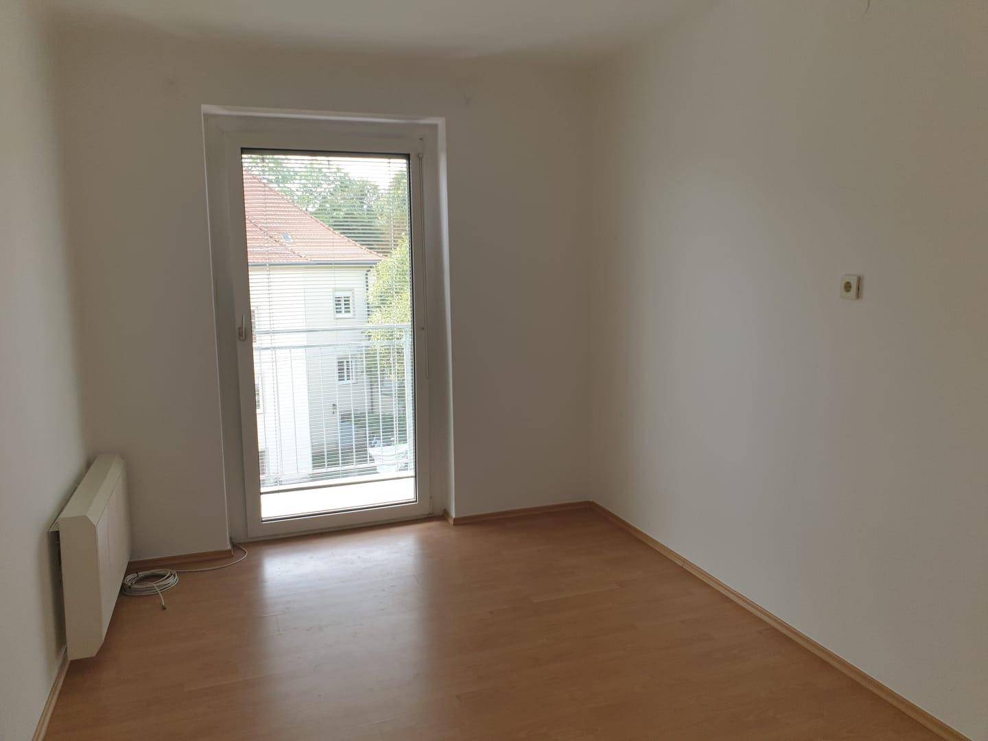 Leeres Zimmer mit Laminatboden und einer großen Fenstertür, die zu einem französischen Balkon führt.