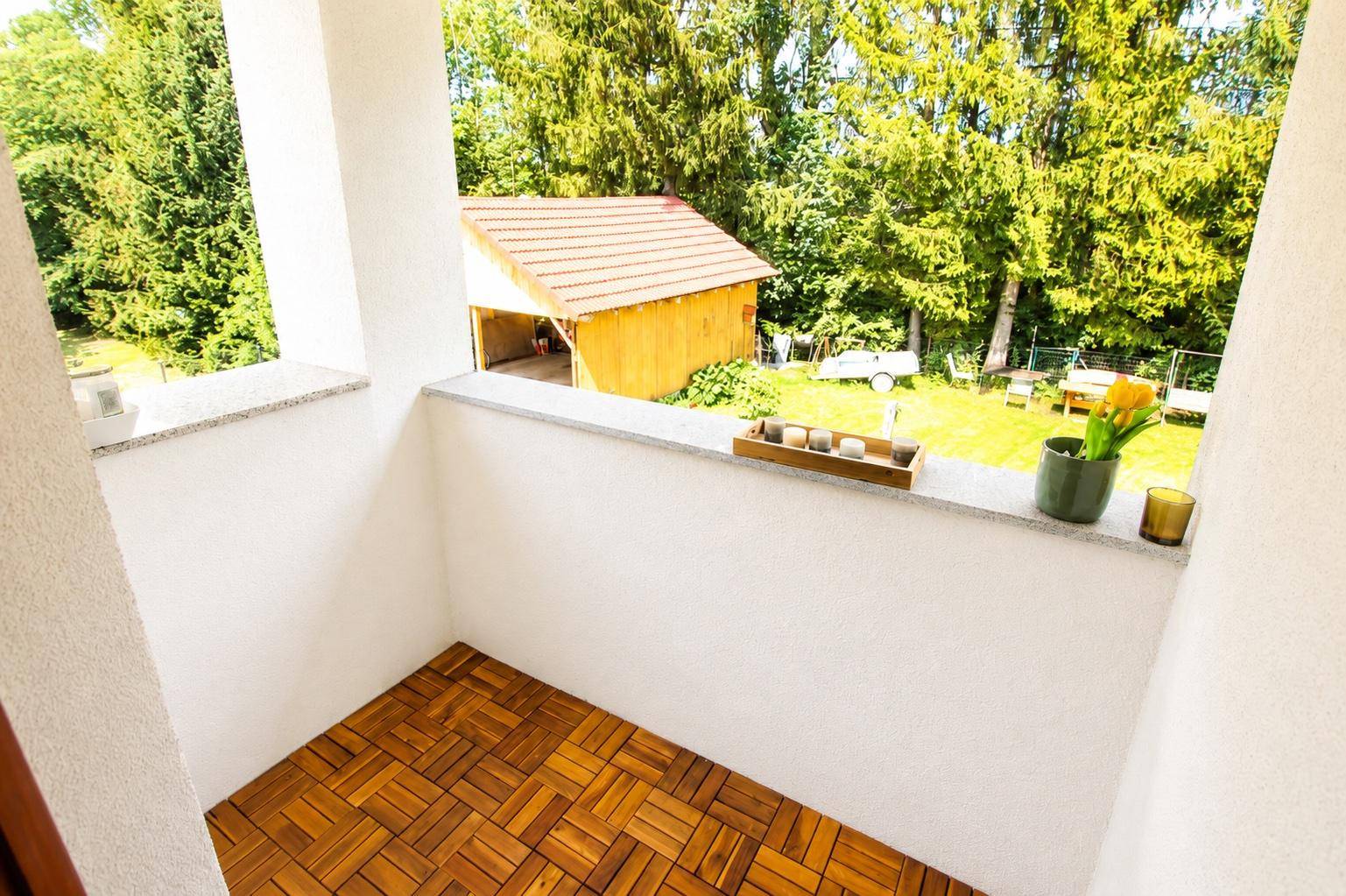 Kleiner Balkon mit Holzbodenfliesen und Blick auf den grünen Garten mit Gartenhaus.