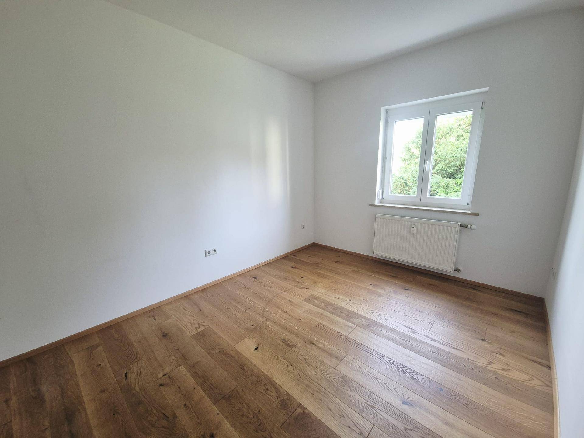 Helles Schlafzimmer mit neuem Eichenparkettboden und einem Fenster mit Blick ins Grüne.