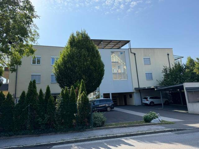 Vorderansicht des Gebäudes mit Eingangsbereich, Bäumen und überdachten Parkplätzen.