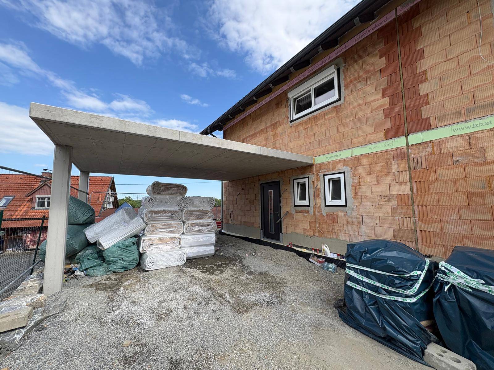Vorderansicht der Doppelhaushälfte im Bau mit Carport und gelagerten Baumaterialien.