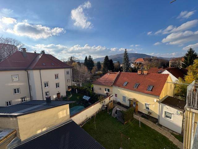 Weitblick über Wohngebäude, Gärten und bewaldete Hügel unter einem klaren blauen Himmel.