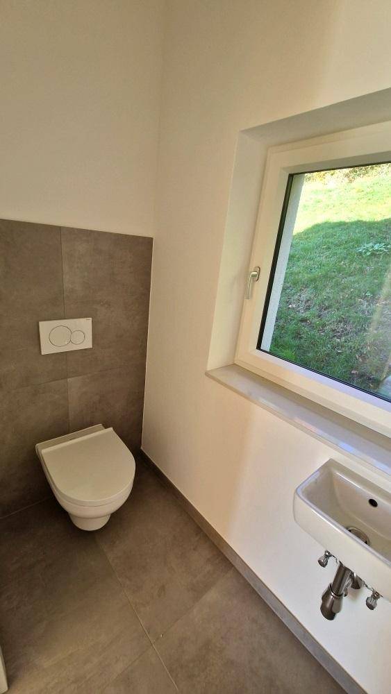 Modernes Gäste-WC mit wandhängender Toilette, kleinem Waschbecken und Fenster mit Blick ins Grüne.