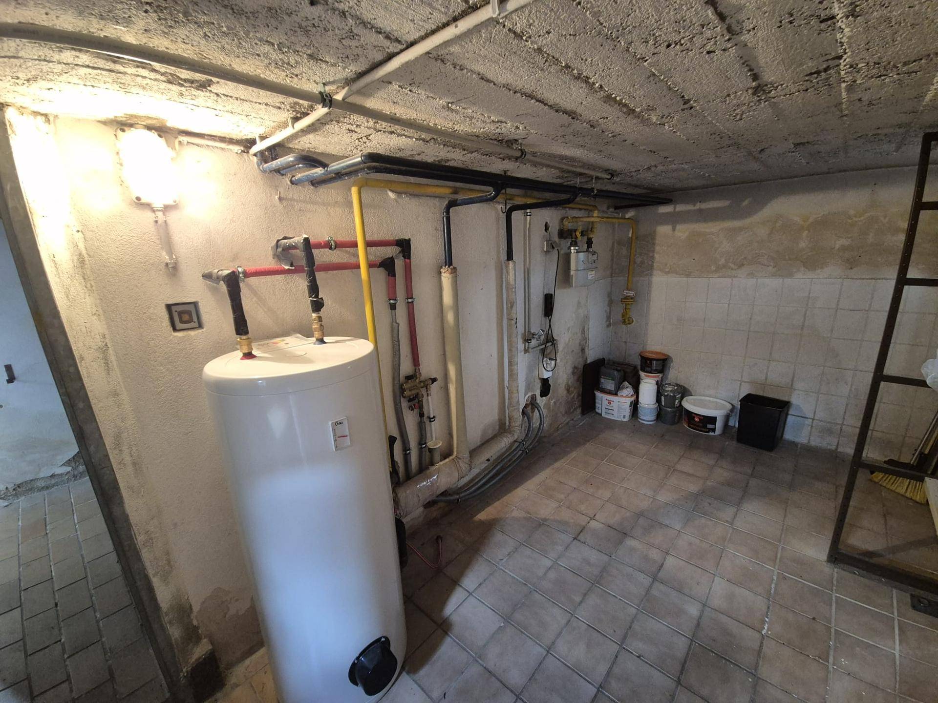 Kellerraum mit Warmwasserbereiter, sichtbaren Leitungen und Fliesenboden.