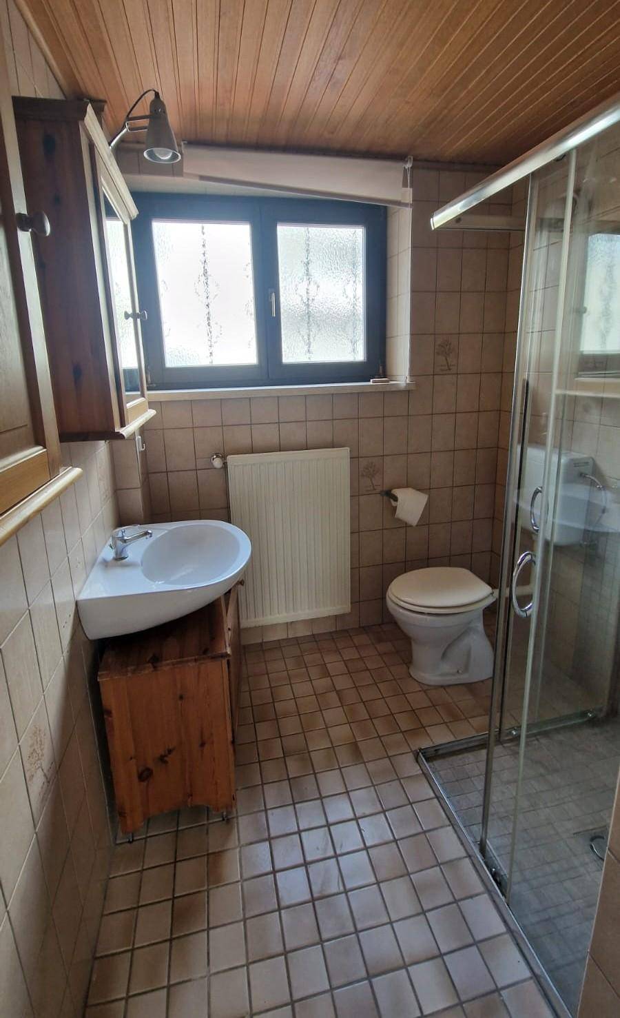 Gefliestes Badezimmer mit Dusche, Waschbecken, Toilette und einem Fenster für Tageslicht.