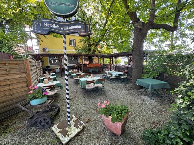 Gemütlicher Gastgarten mit Tischen unter schattenspendenden Bäumen und Willkommensschild.