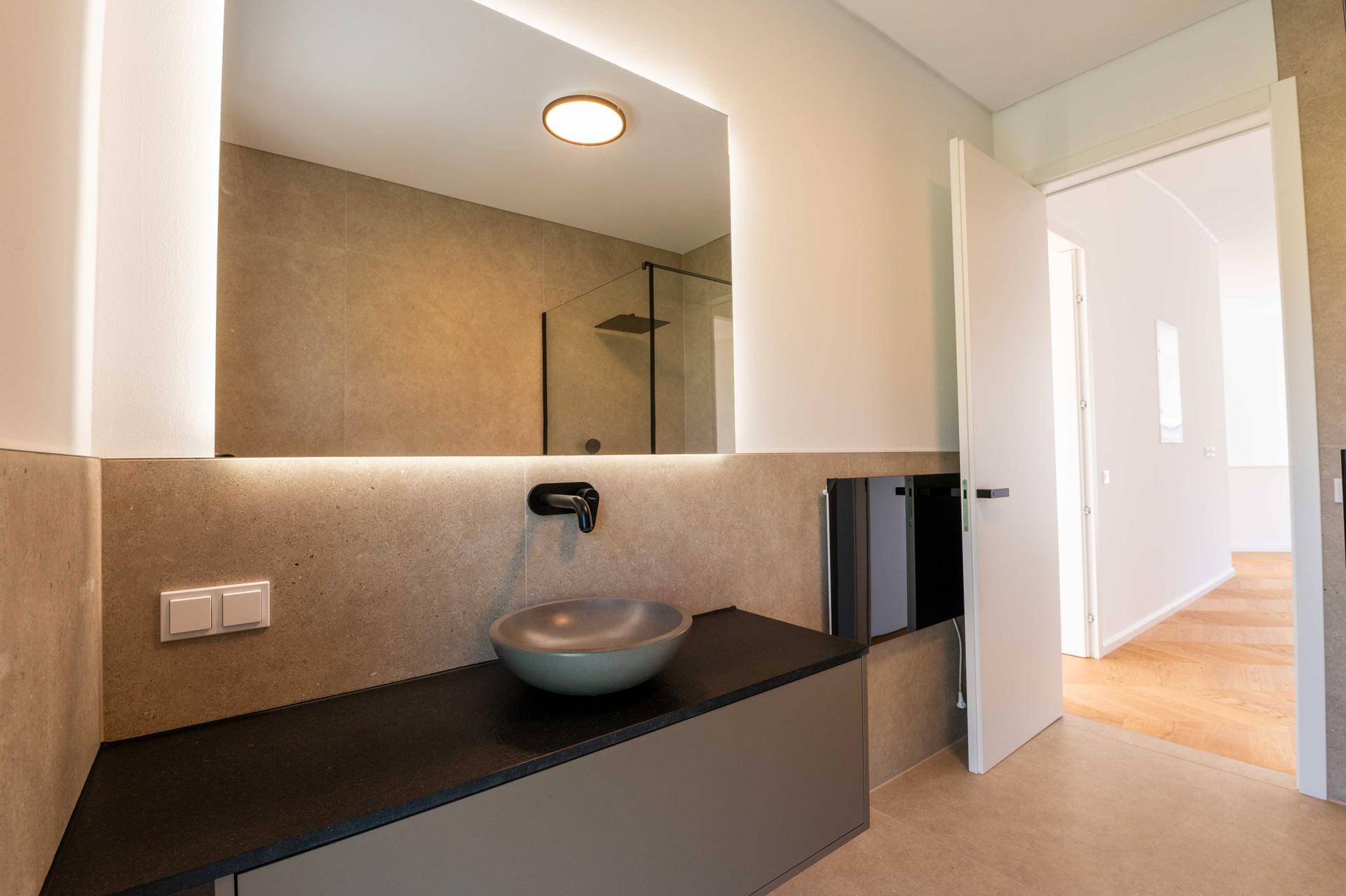 Modernes Badezimmer mit beleuchtetem Spiegel, Waschtisch und Zugang zum Schlafzimmer.