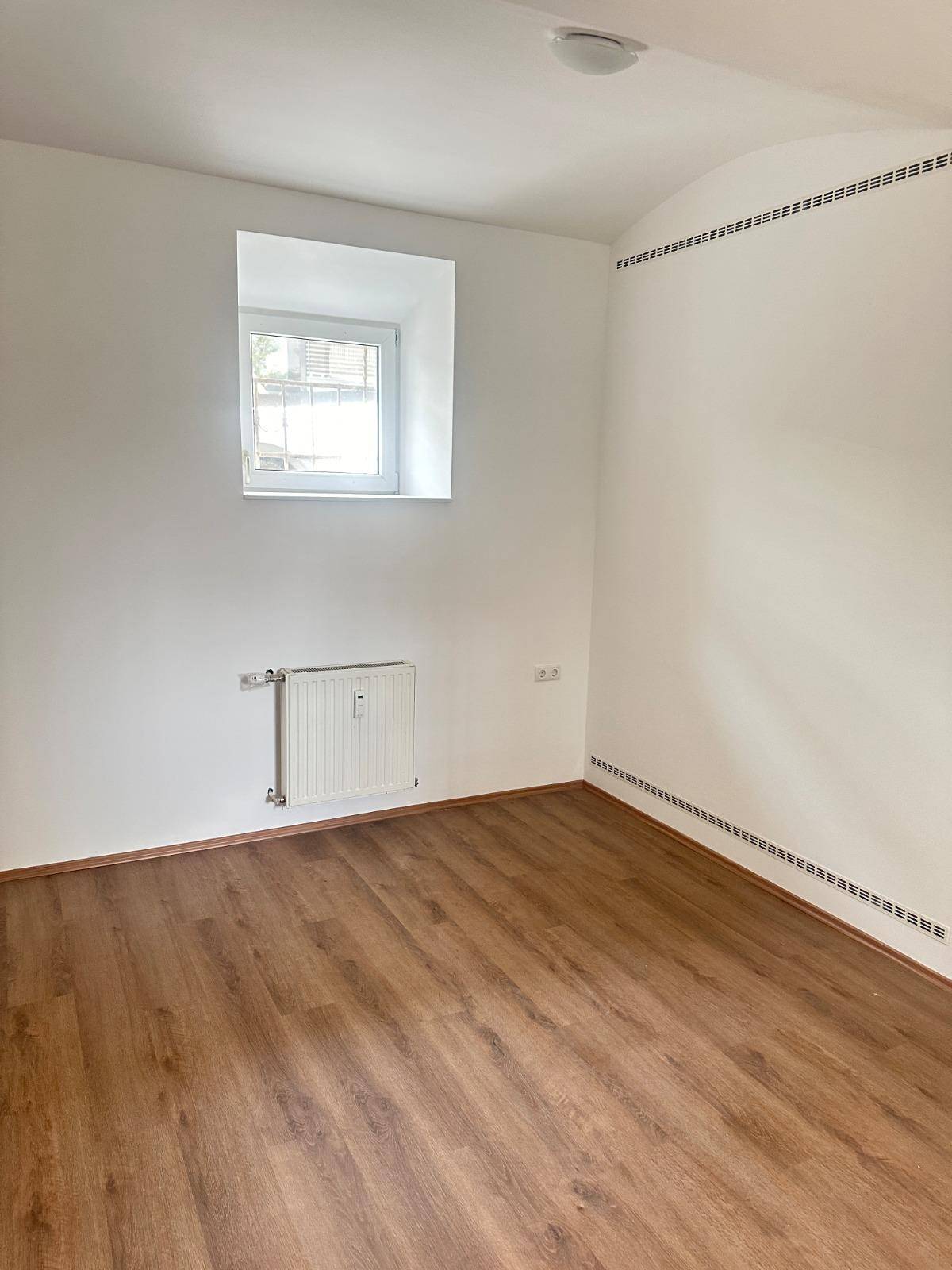 Helles Schlafzimmer mit Laminatboden und einem kleinen Fenster, das Tageslicht spendet.