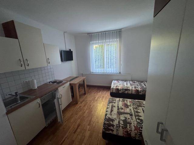 Studio-Apartment mit zwei Betten, kleiner Küchenzeile und Fenster mit Jalousien.